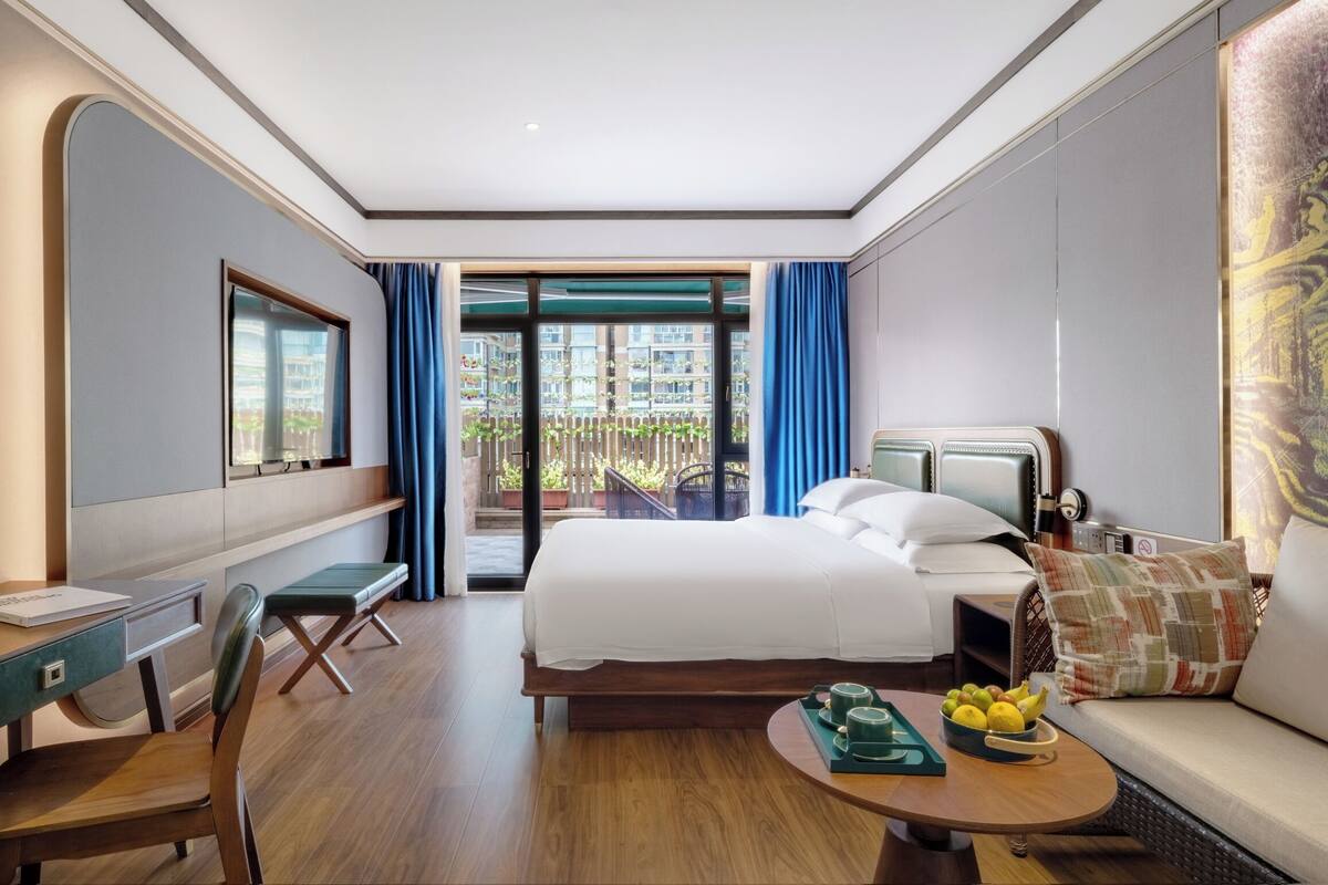 Une chambre d’hôtel avec un lit, un bureau, une chaise, un canapé, une table basse avec des fruits, et une vue sur le balcon extérieur.