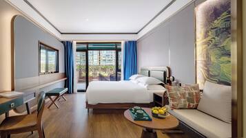 A hotel room with a bed, a desk, a chair, a sofa, a coffee table with fruits, and a view of the outside balcony.