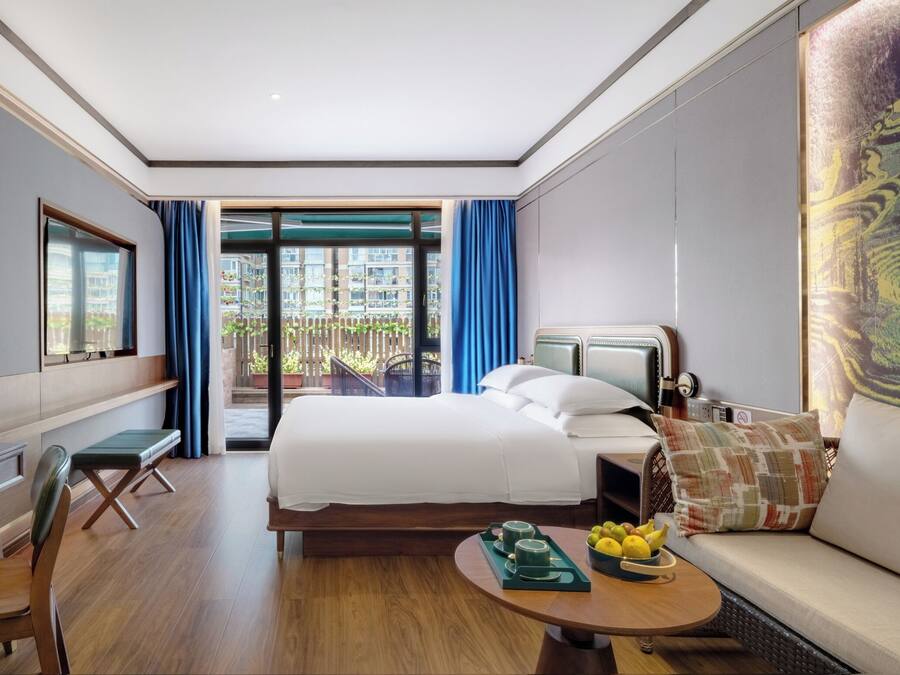 A hotel room with a bed, a desk, a chair, a sofa, a coffee table with fruits, and a view of the outside balcony.