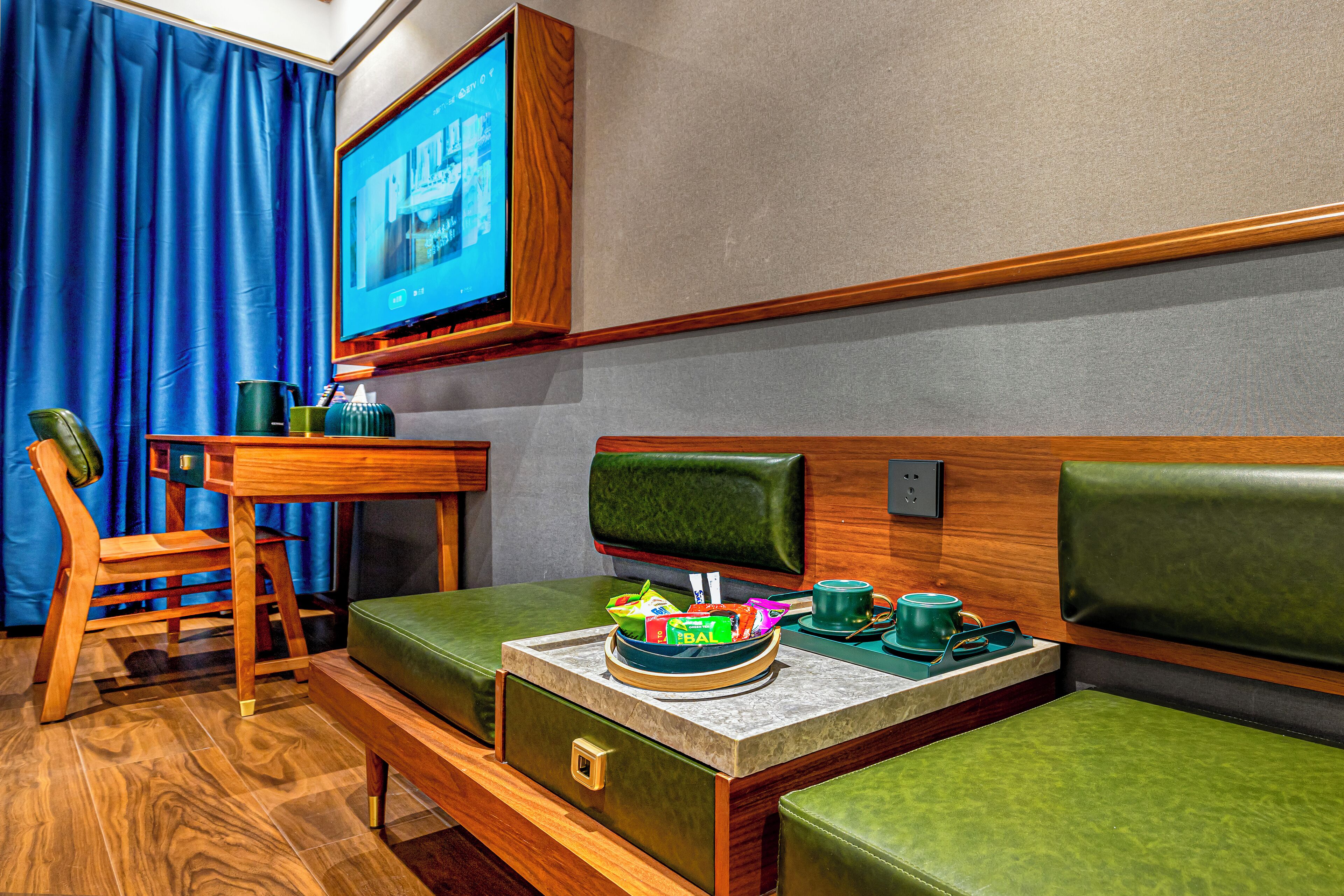 A compact room with a wooden desk, a green cushioned bench, a television mounted on the wall, and a small table with a tray of snacks.