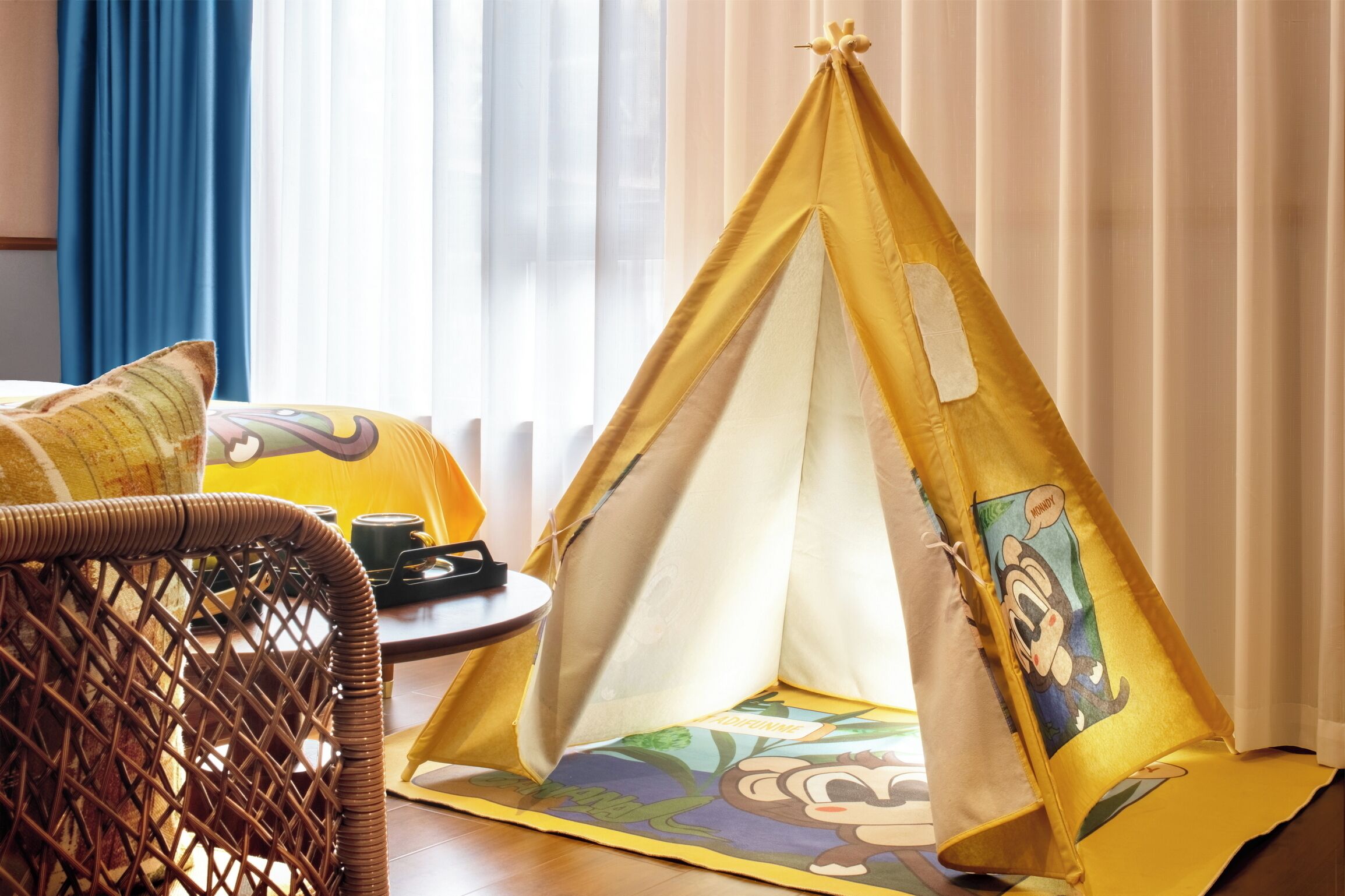 A room with a yellow tent-like structure featuring cartoon characters, a wicker chair, and a small table with a cup and a remote control.