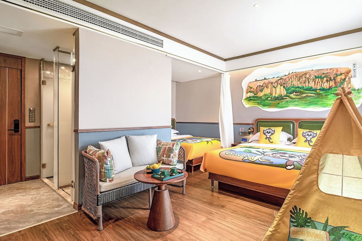 Une chambre d’hôtel avec une armoire en bois, un miroir rond, un lit avec une literie aux motifs de dessins animés, une petite table avec des fruits et une décoration évoquant une tente.