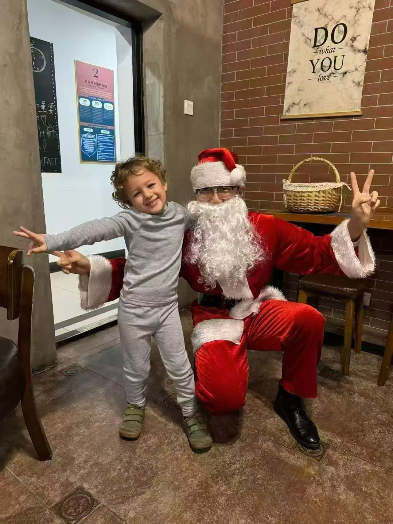 A child and a person in a Santa costume sitting on a bench in a room with a brick wall and a poster that says "DO what YOU love".