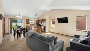 A spacious living area with a gray sofa, a dining table with chairs, a kitchen with modern appliances, and a flat-screen TV mounted on the wall.