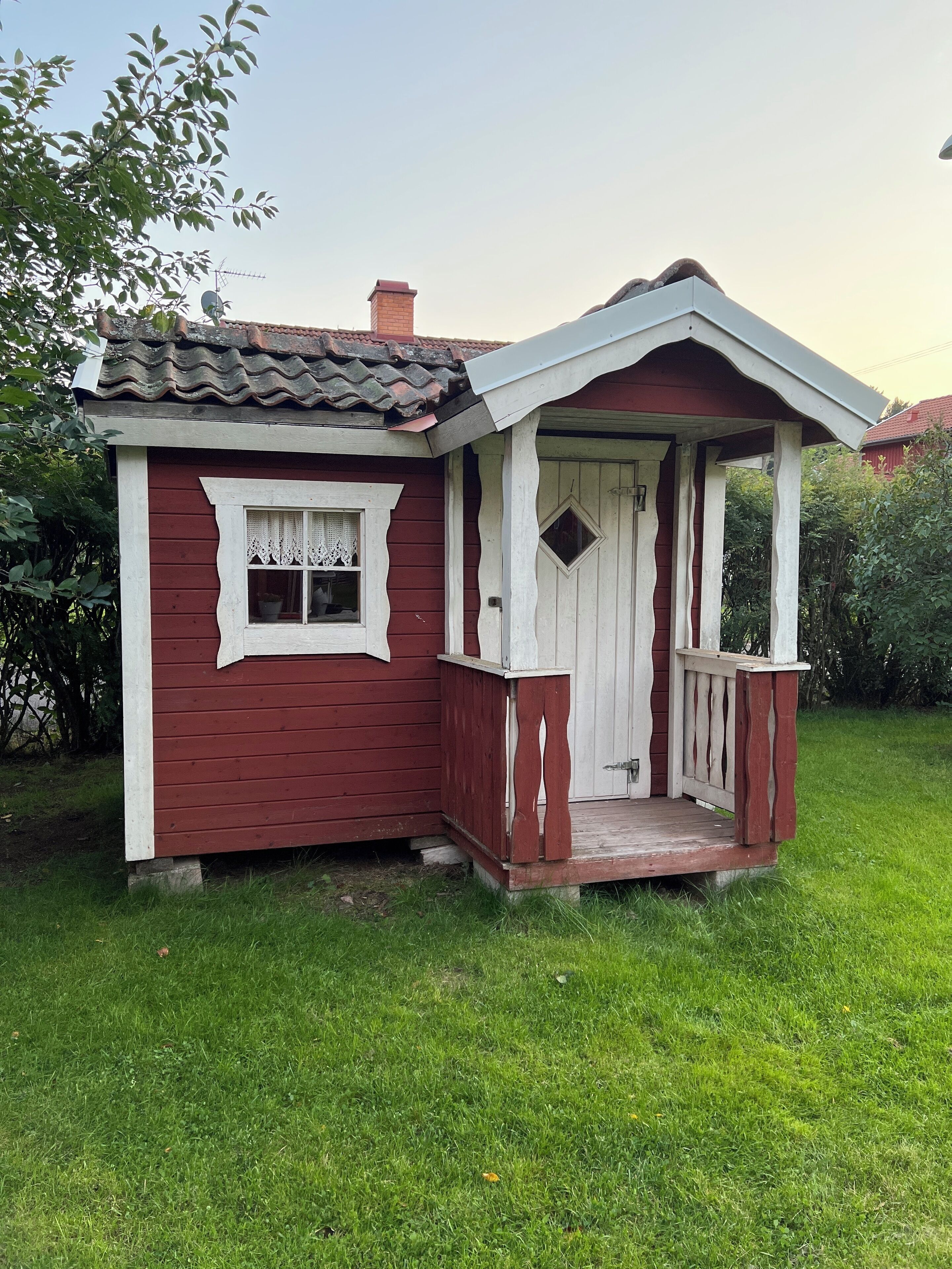 Een klein, gelijkvloers huis met een rode gevel en witte details, met een veranda met een witte balustrade en een deur met een ruitvormig raam.