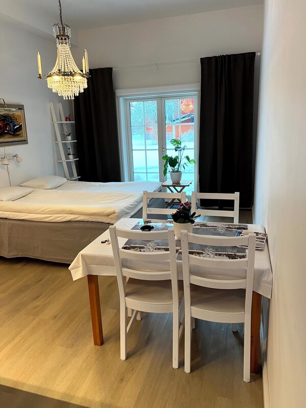A bedroom with a bed, a table with chairs, a chandelier, a window with curtains, and a plant. - Flygeln lägenhetshotell (Leksand)