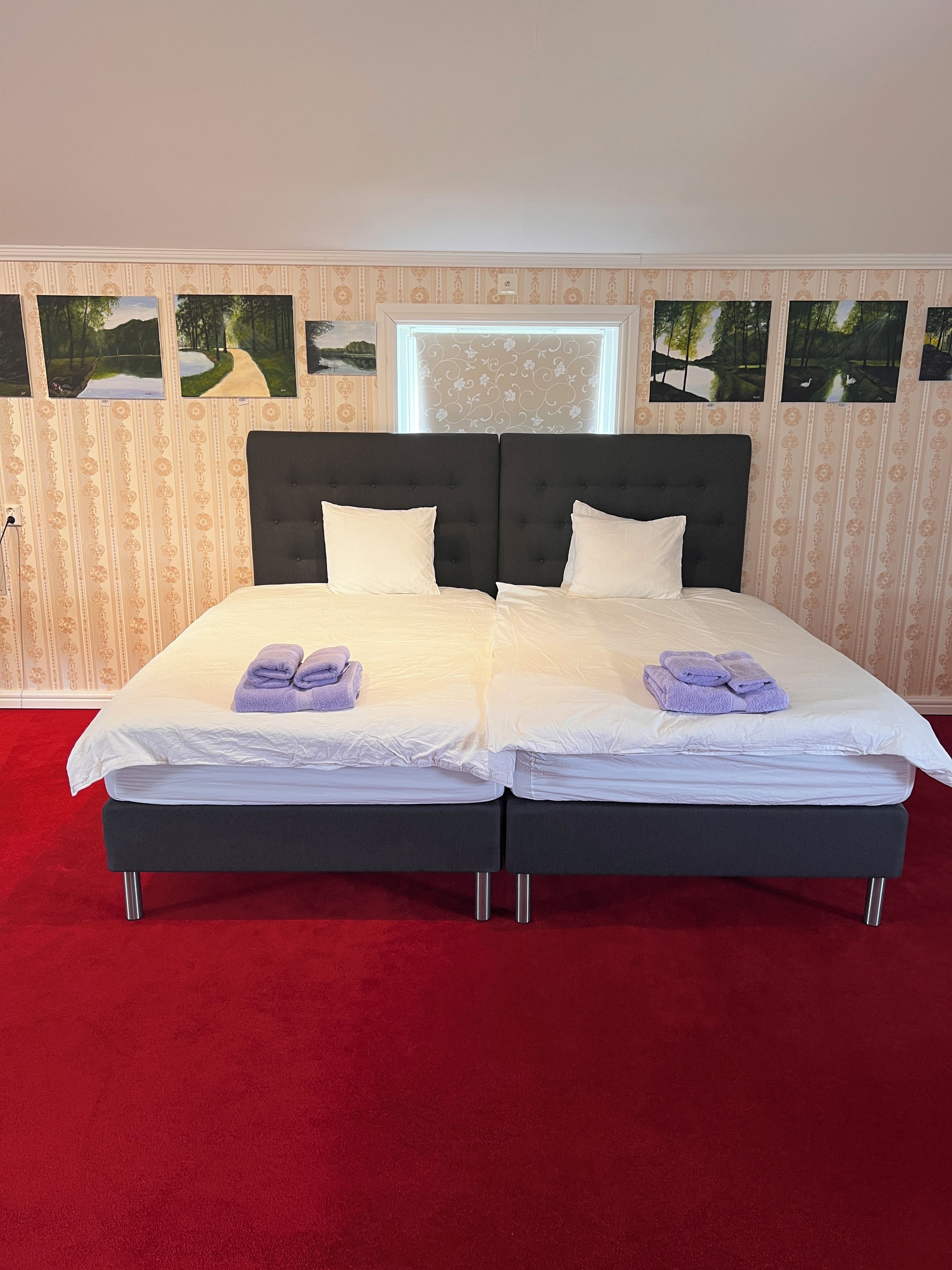 Cama doble con sábanas blancas y dos toallas moradas, sobre una alfombra roja en una habitación con papel tapiz estampado y cuadros en la pared.