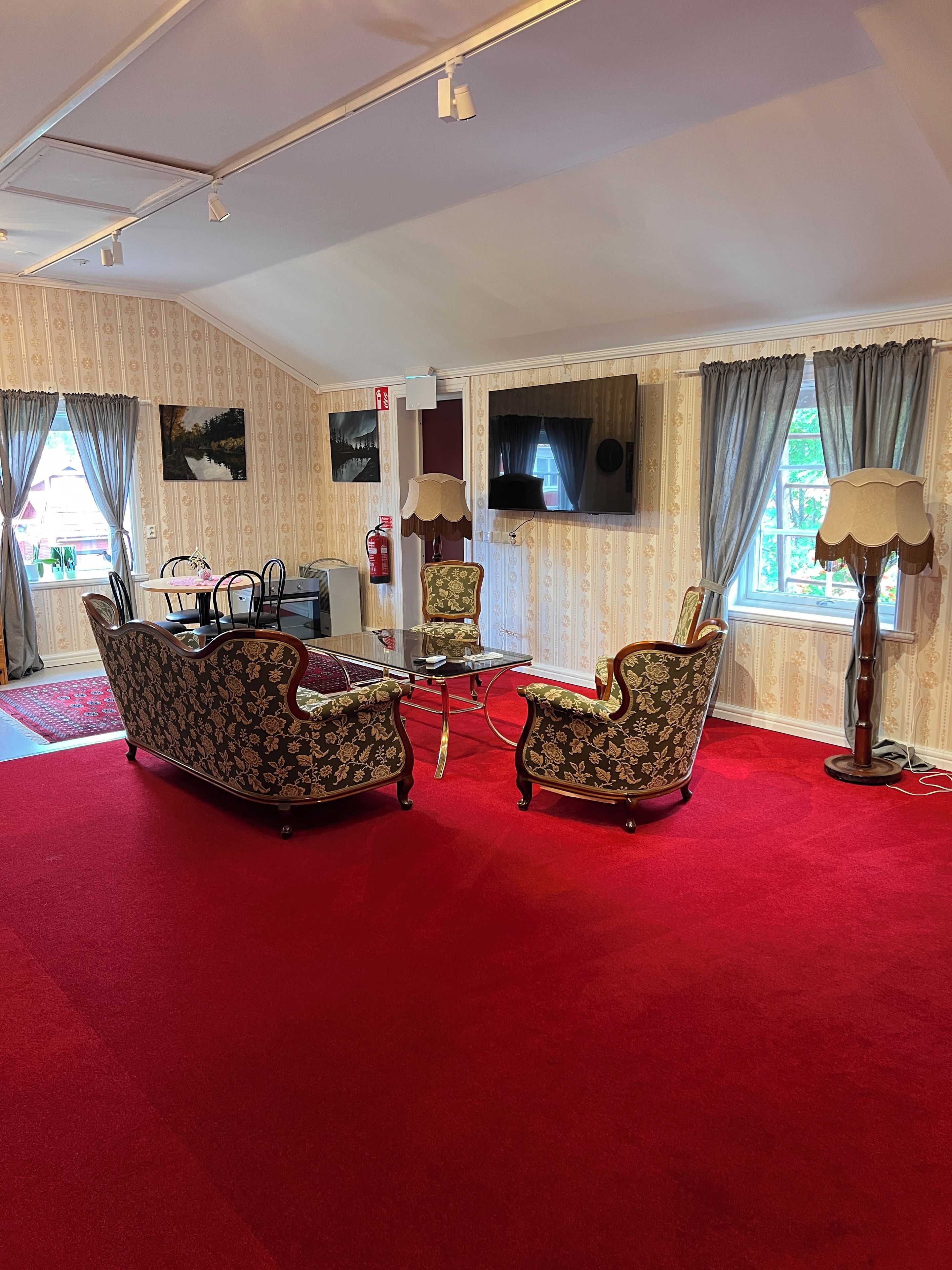 Una habitación con alfombra roja, dos sillones, una mesita, un televisor y una ventana con cortinas.