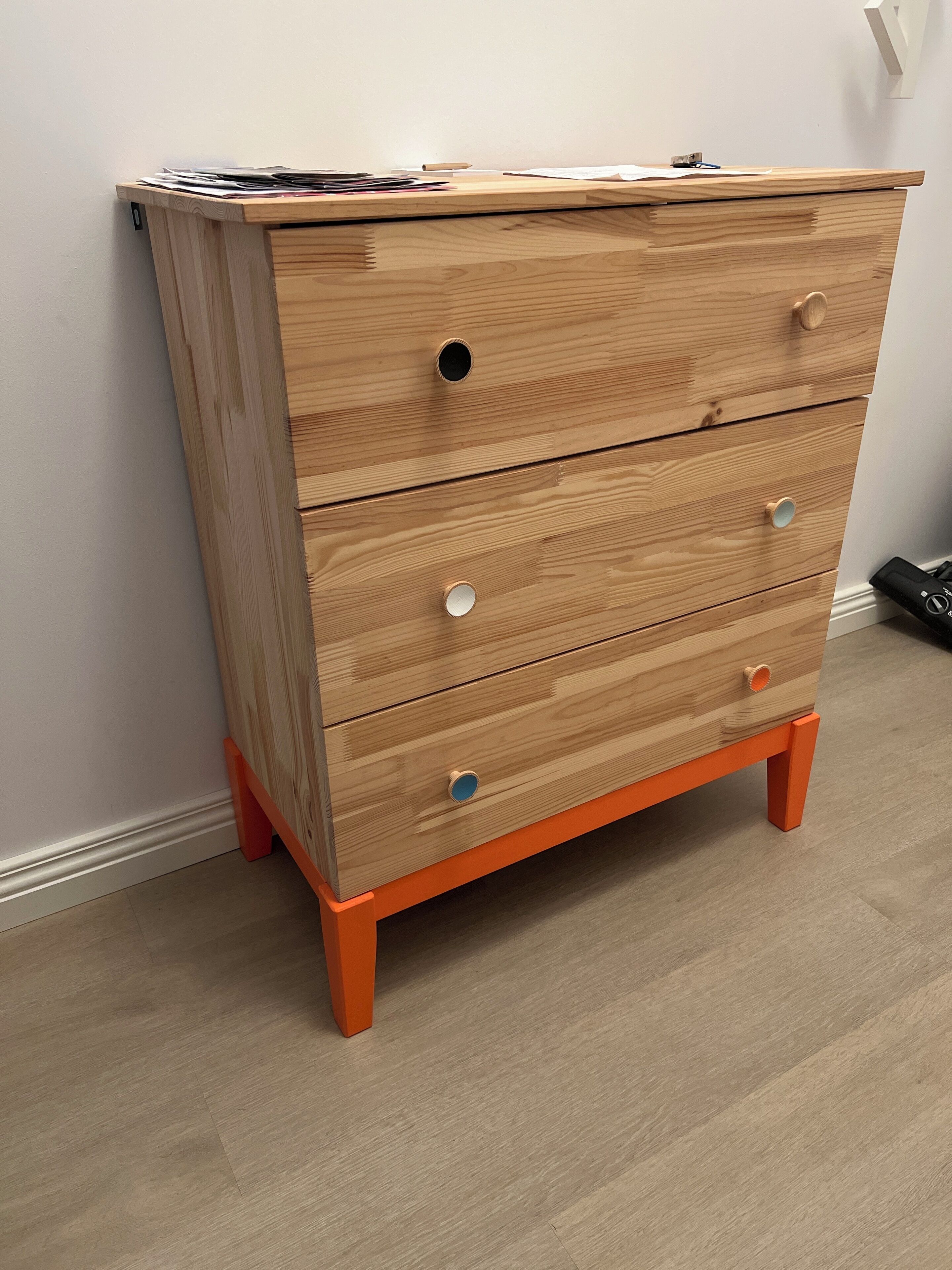 Un aparador de madera con tres cajones y un estante superior, patas color naranja y una pared blanca de fondo.