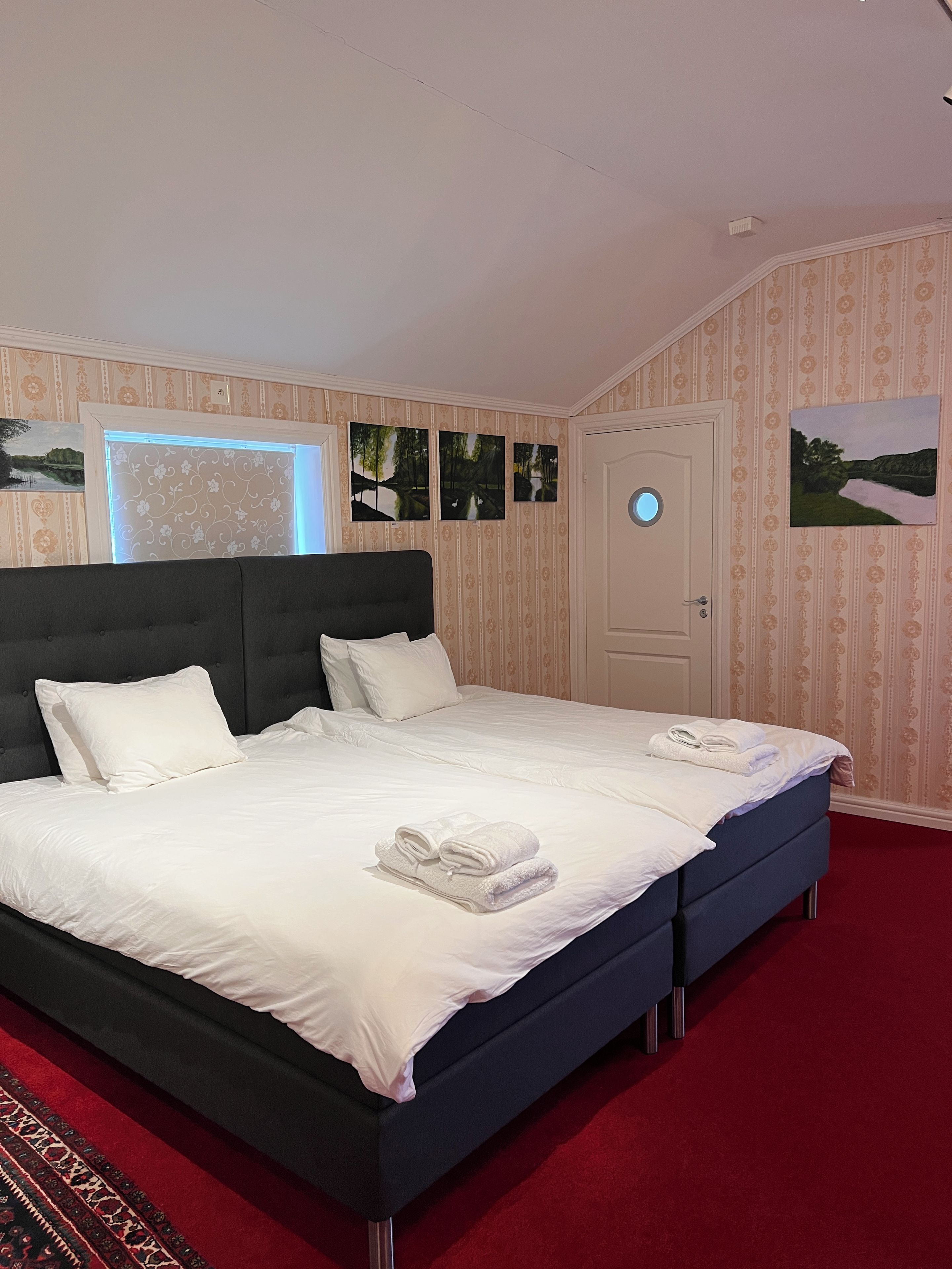 A hotel room with a large bed, a door with a round window, and a red carpet.