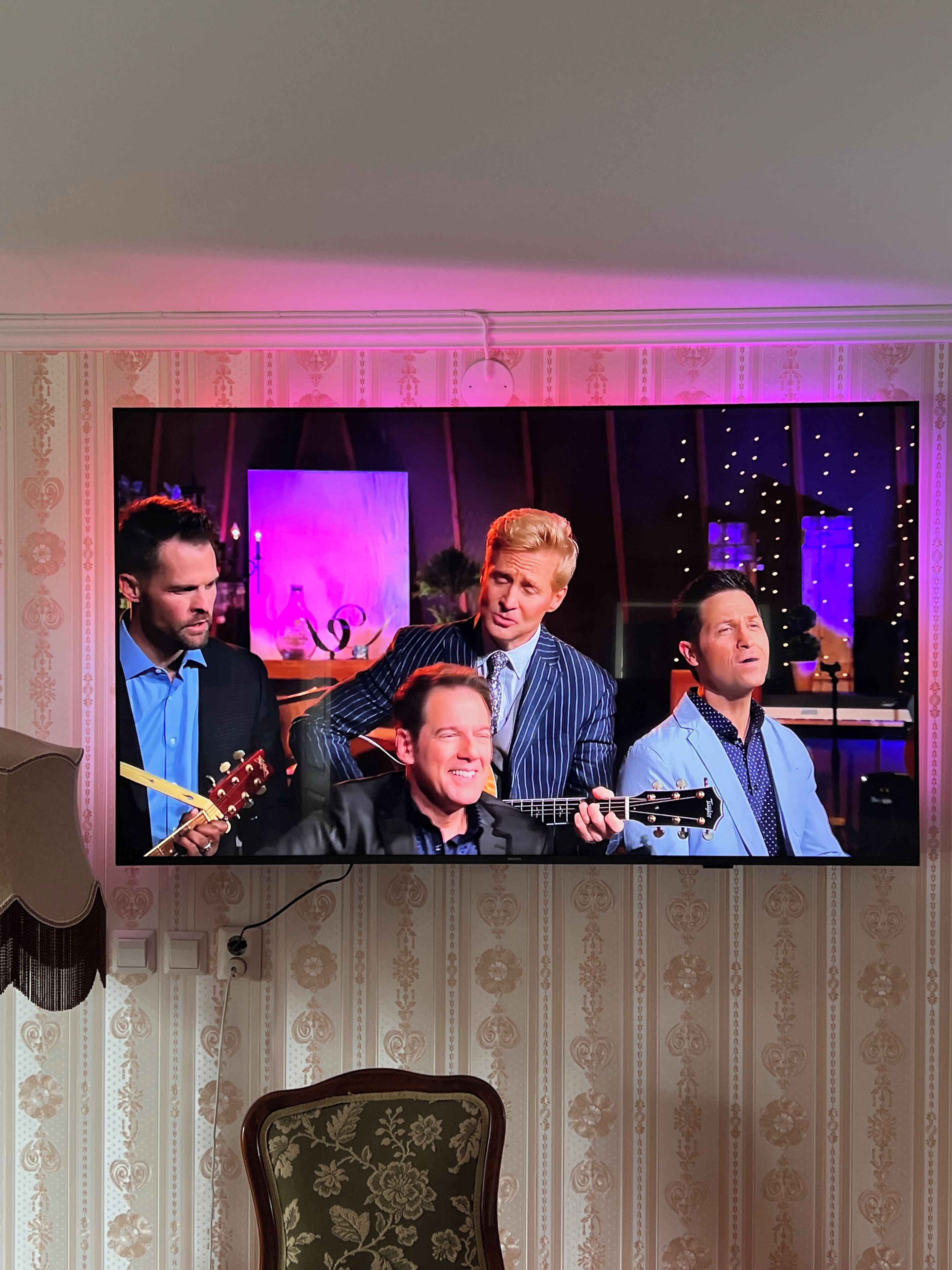 A television displaying a band performing, with a patterned wallpaper and a chair in the foreground.