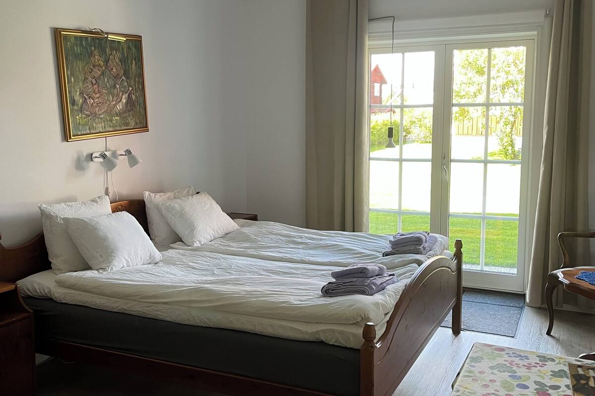 A bedroom with a large bed, a bedside table, a lamp, a painting on the wall, and a view of the outdoors through a window.