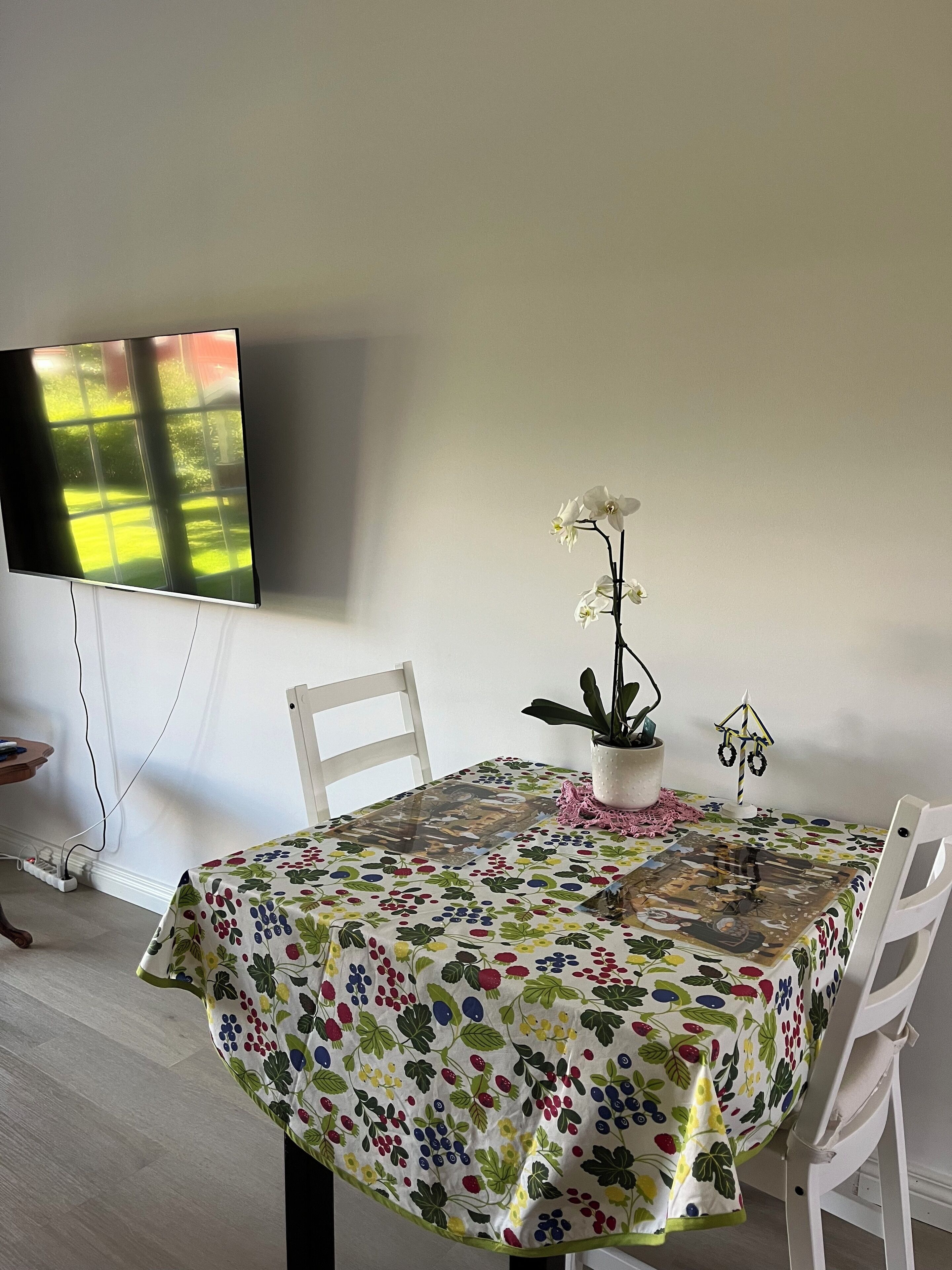 A dining area with a table covered in a floral tablecloth, chairs, a potted orchid, and a wall-mounted television.