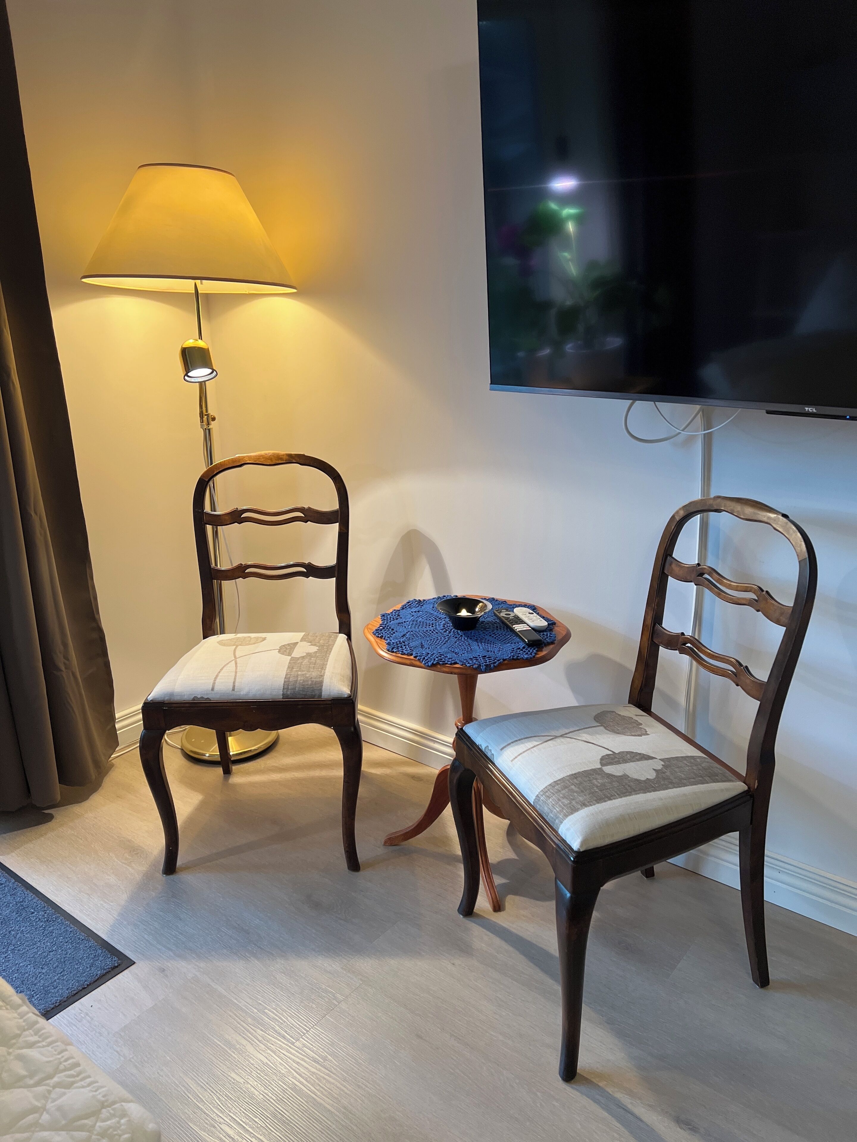 A hotel room with two wooden chairs, a small round table with a blue table runner, a lamp, and a flat-screen TV mounted on the wall.