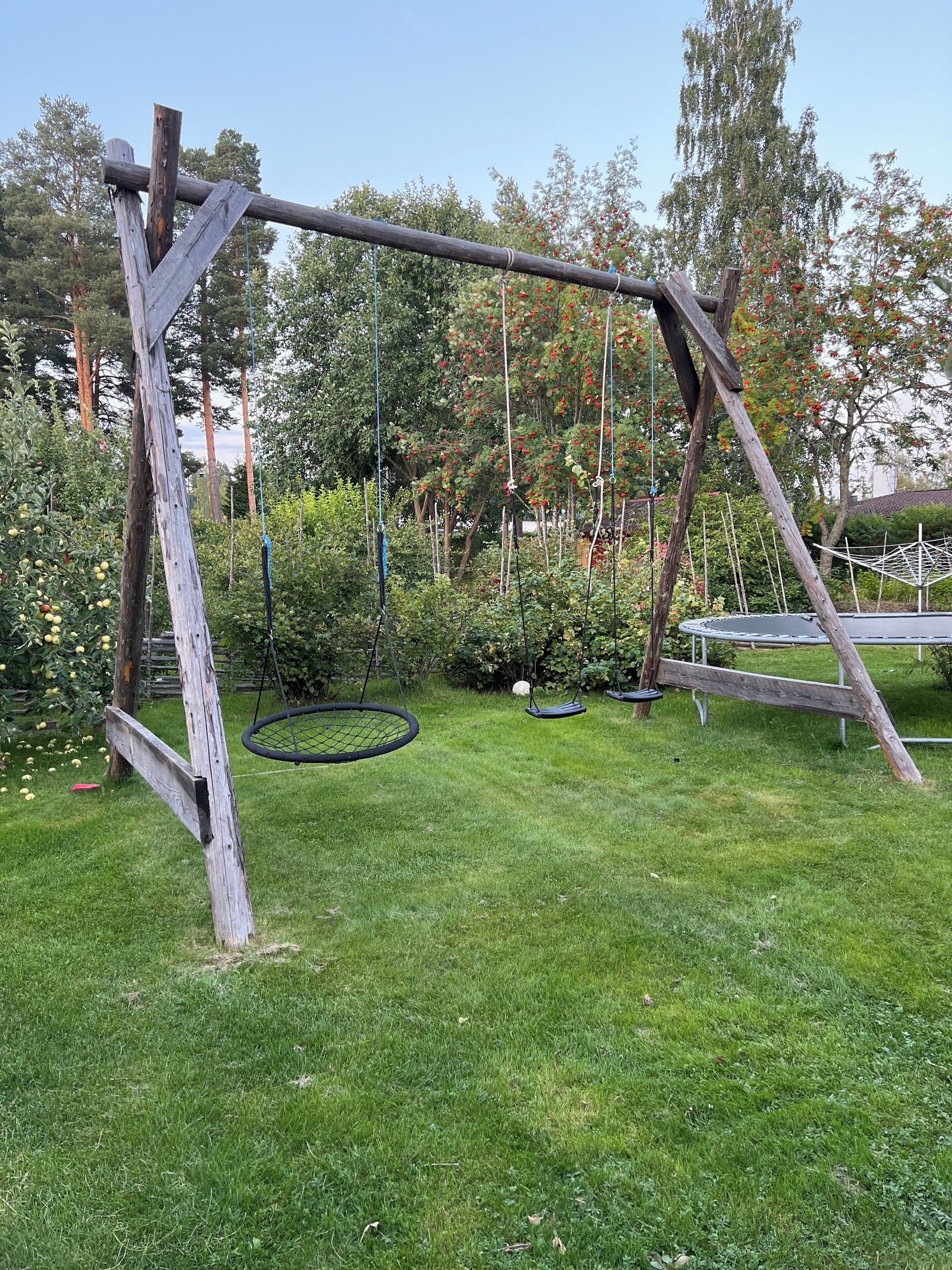 A wooden swing set in a grassy yard with trees and a trampoline in the background.