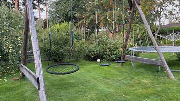 A wooden swing set in a grassy yard with trees and a trampoline in the background.
