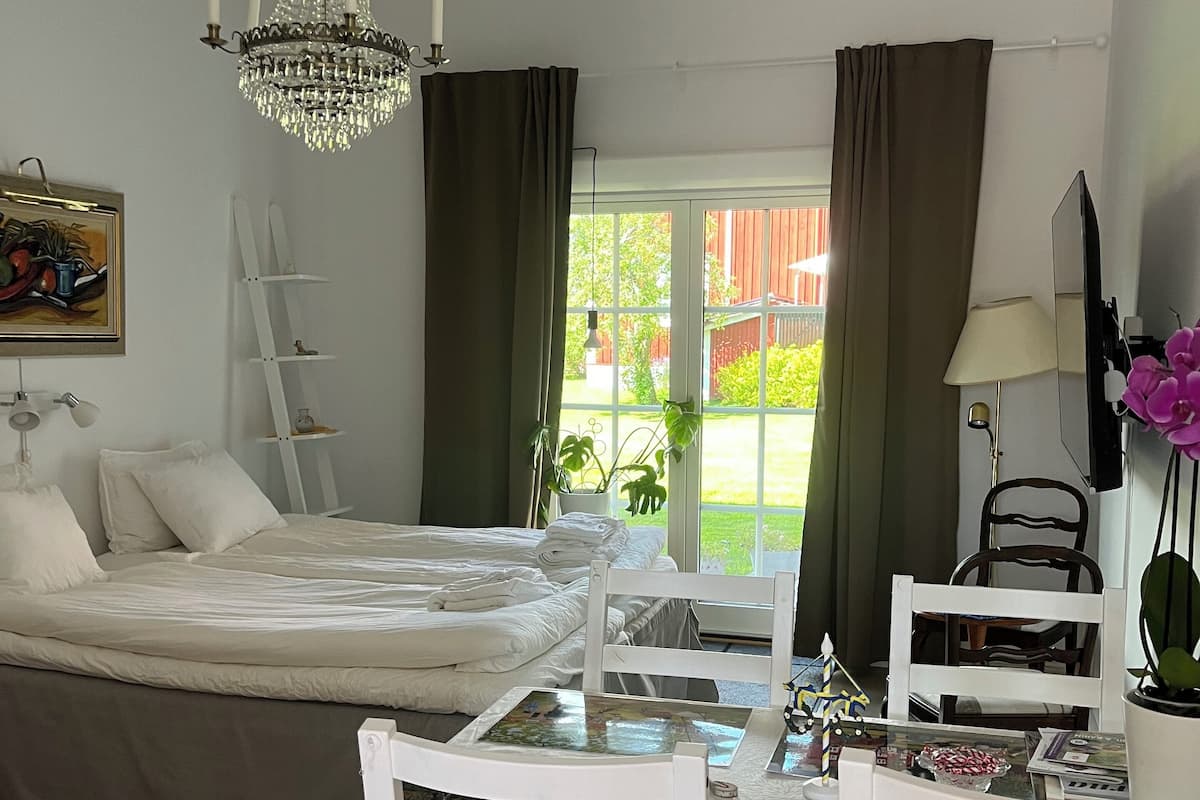 A bedroom with a bed, a chandelier, a window with curtains, a desk with chairs, and a painting on the wall.