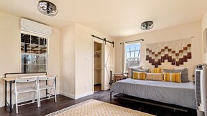 Een slaapkamer met een bed, nachtkastje en een bureau met stoelen. Er is een raam met uitzicht, een plafondventilator en een airconditioner.