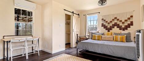 A bedroom with a bed, bedside table, and a desk with chairs. There is a window with a view, a ceiling fan, and an air conditioning unit.