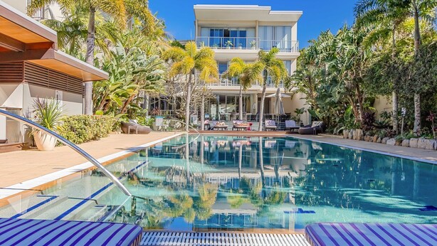 Piscina com espreguiçadeiras, cercada por plantas tropicais e um edifício moderno ao fundo.