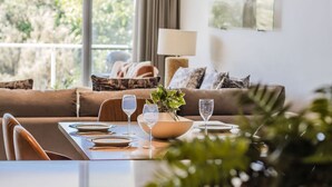 Uma sala de jantar com mesa posta para dois, planta em vaso e vista para uma varanda com sofá e almofadas.