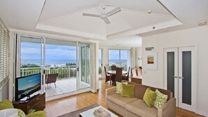 A spacious living room with a large TV, a sofa with cushions, a coffee table, and a balcony with a view of the ocean.