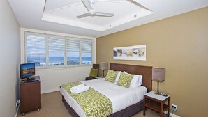 A hotel room with a bed, a desk with a computer, a ceiling fan, a window with blinds, and a wall-mounted artwork.