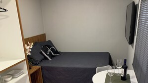 Quarto compacto com cama, uma pequena mesa com duas taças de vinho e uma garrafa, uma cadeira, prateleira com pratos e um tapete macio.
