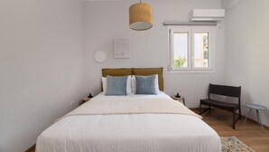 A bedroom with a bed, a wooden bench, a small table, and a window with an air conditioner.