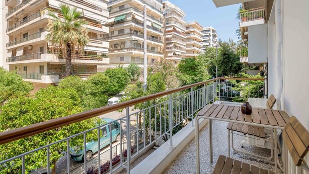A balcony with a wooden table and chairs, overlooking a parking area and a row of multi-story buildings with balconies.