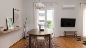 A modern living room with a round wooden dining table, white chairs, a large flat-screen TV, and a ceiling-mounted air conditioner.