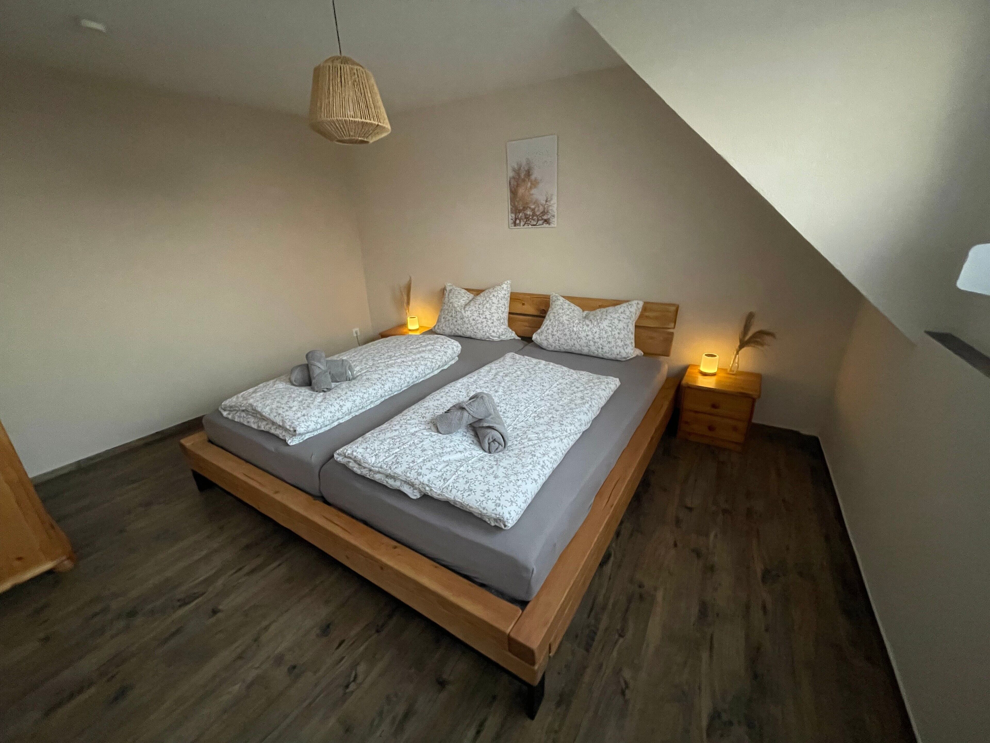 A bedroom with a wooden bed, two bedside tables, a hanging light fixture, and a wall-mounted picture.