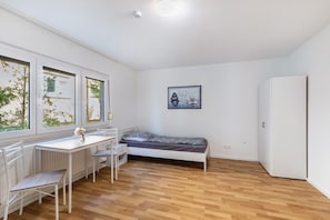 Ein kleiner, heller Raum mit einem Bett, einem Tisch und Stühlen, großen Fenstern und einer Heizung.
