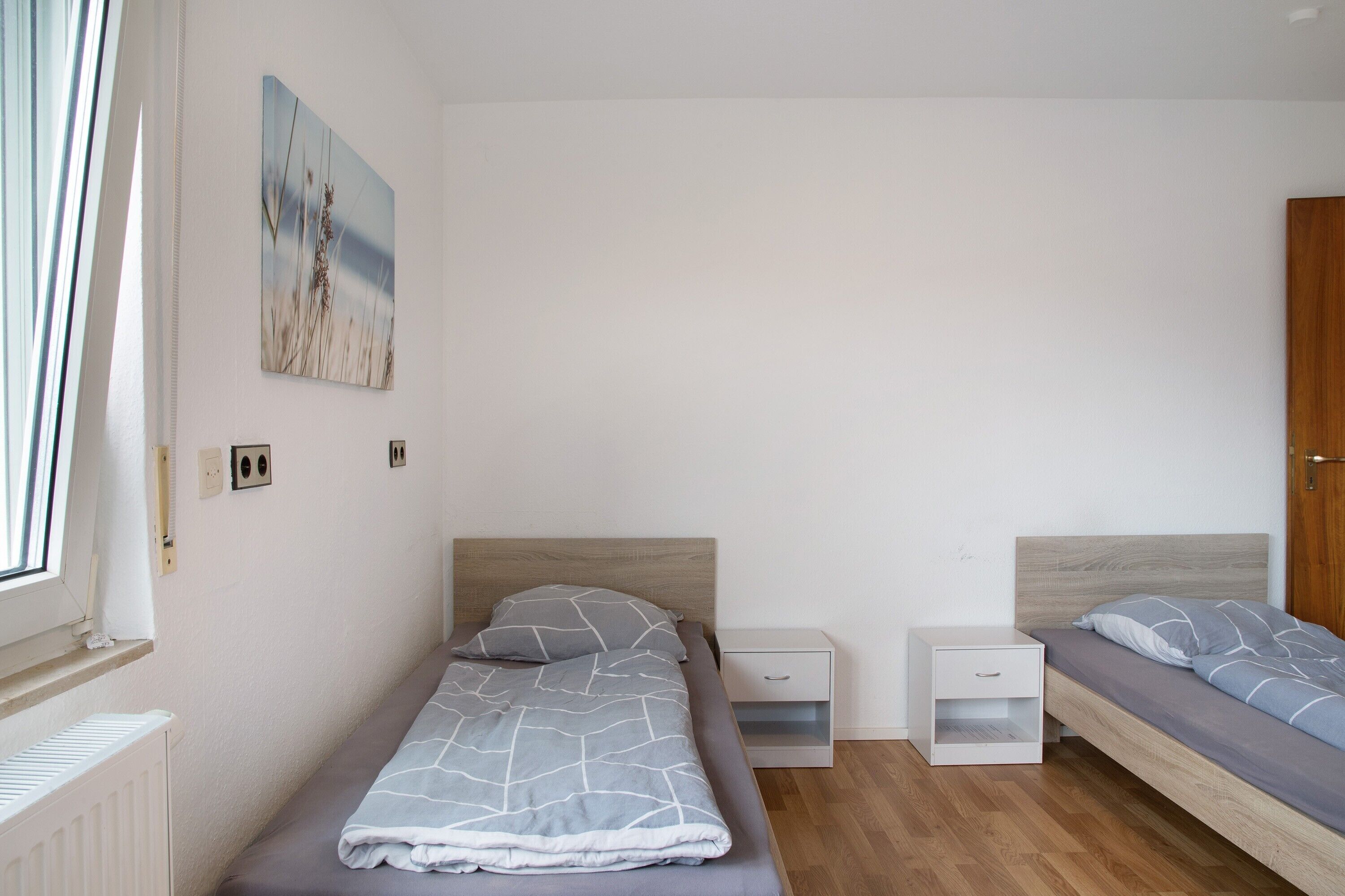 A bedroom with a single bed, a nightstand, a drawer, a wooden headboard, a window, and a door.