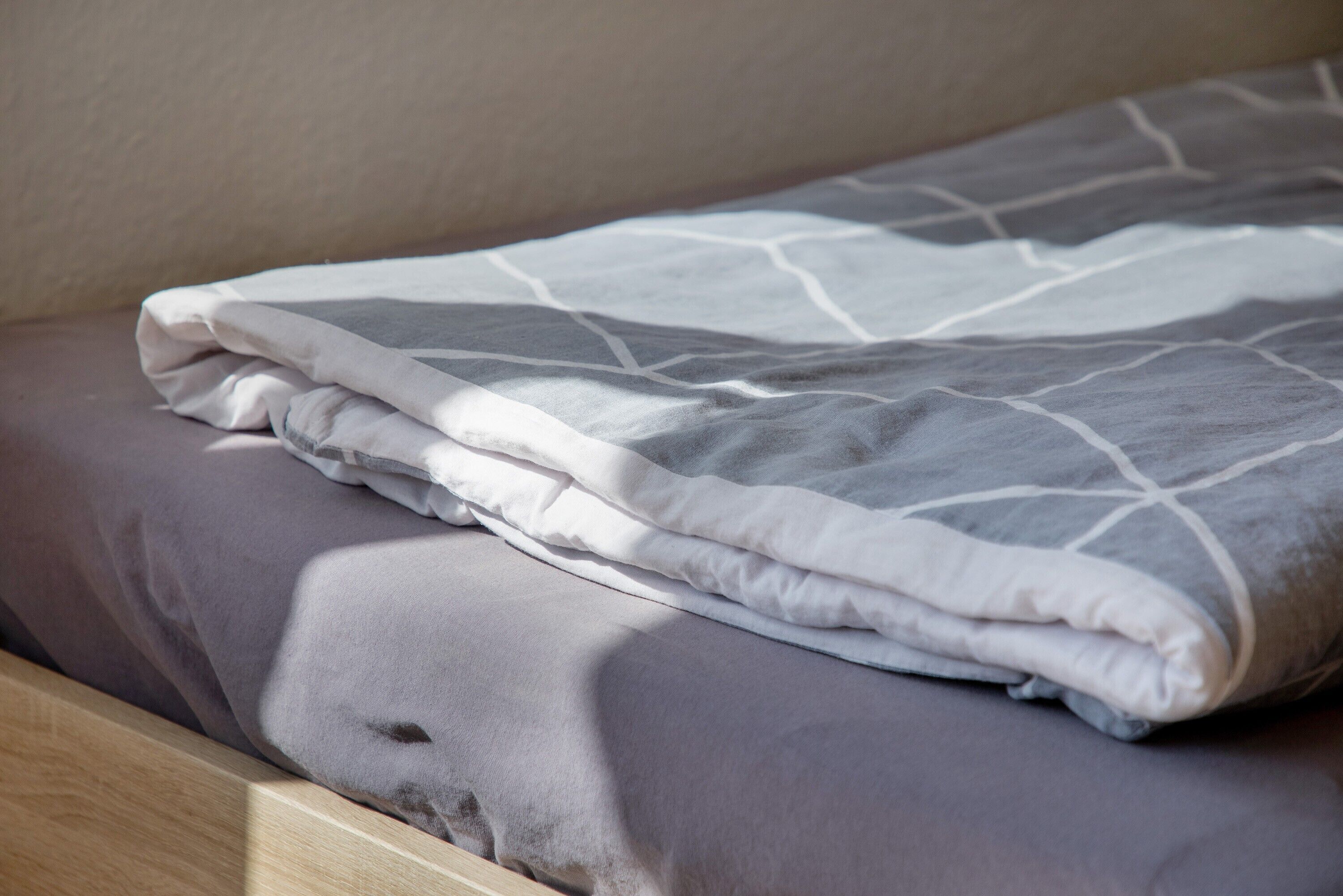 A neatly made bed with a gray and white patterned blanket, a wooden headboard, and a plain wall.