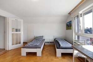 Una habitación pequeña y luminosa con dos camas individuales, piso de madera, ventana con vistas y puerta con paneles de vidrio satinado.