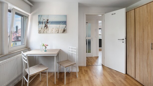 A room with a wooden floor, a white table with two chairs, a painting on the wall, and a door with a number. - Apartment 'Wohnung 06' with Wi-Fi (Waiblingen)