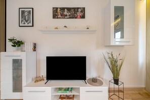En stue med et fladskærms-tv, en reol med pynteting, en potteplante og et billede i ramme på væggen.