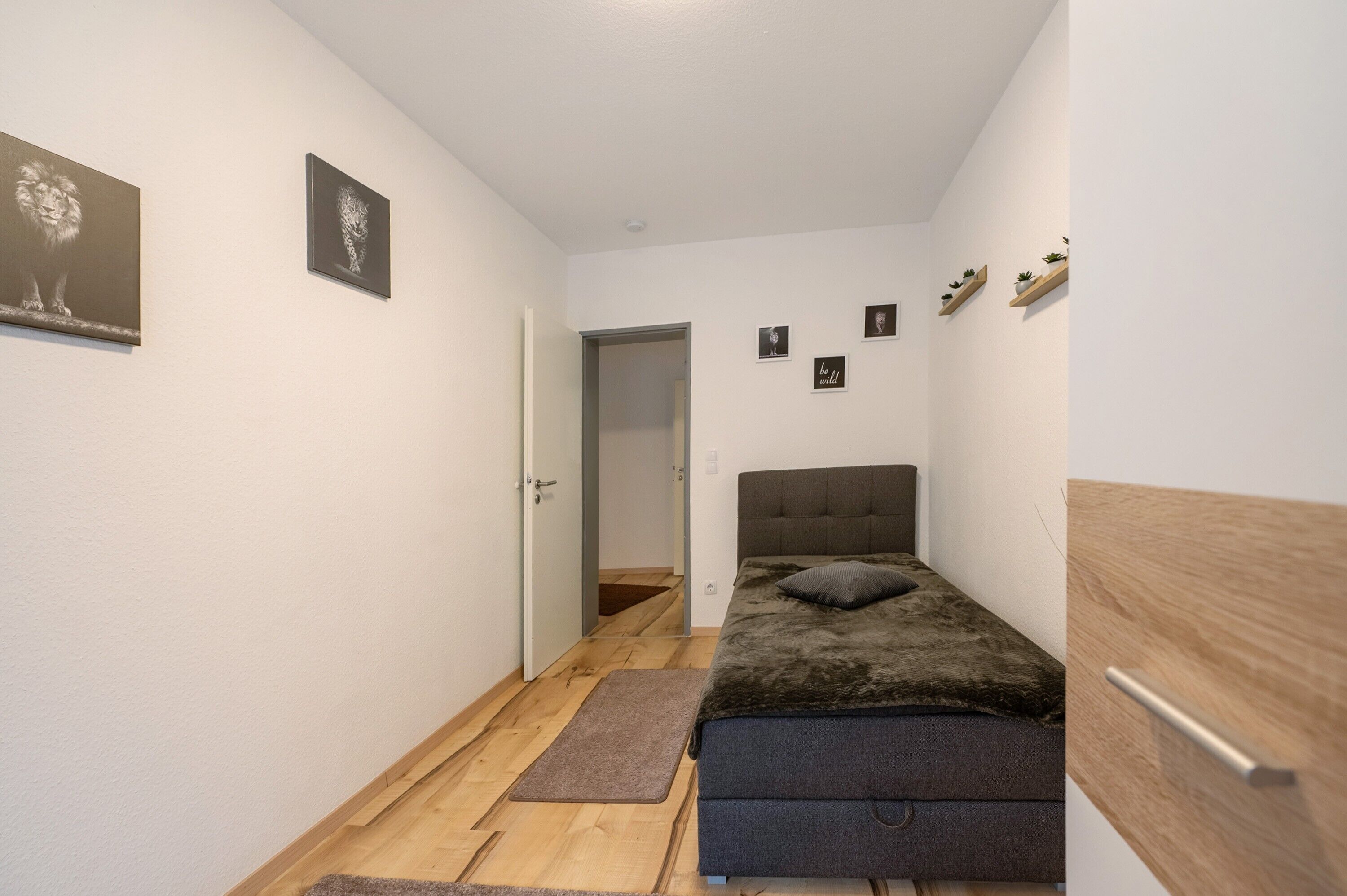Una habitación pequeña y estrecha con una cama, piso de madera y una puerta que da a otra habitación.