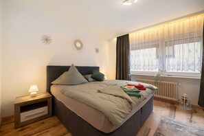 Et soveværelse med seng, sengebord, lampe, vindue med gardiner, radiator og dekorative dekorationer på væggen.
