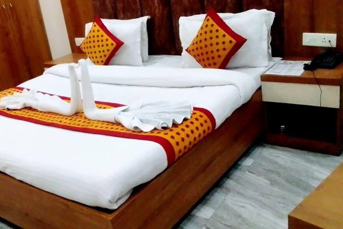 A neatly made bed with white linens, a red and yellow bedspread, and decorative pillows. There is a wooden nightstand with a phone and a small table beside the bed.