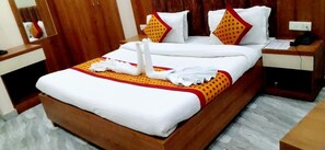 A neatly made bed with white linens, a red and yellow bedspread, and decorative pillows. There is a wooden nightstand with a phone and a small table beside the bed. - Hotel Daksh (Haridwar)