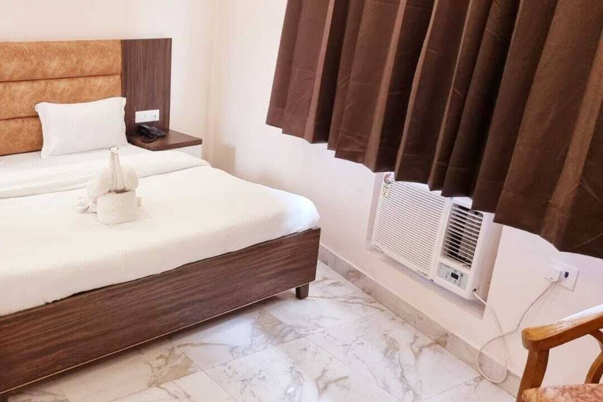 A neatly made bed with white linens and a decorative pillow, a bedside table with a lamp, and a wall-mounted air conditioner.