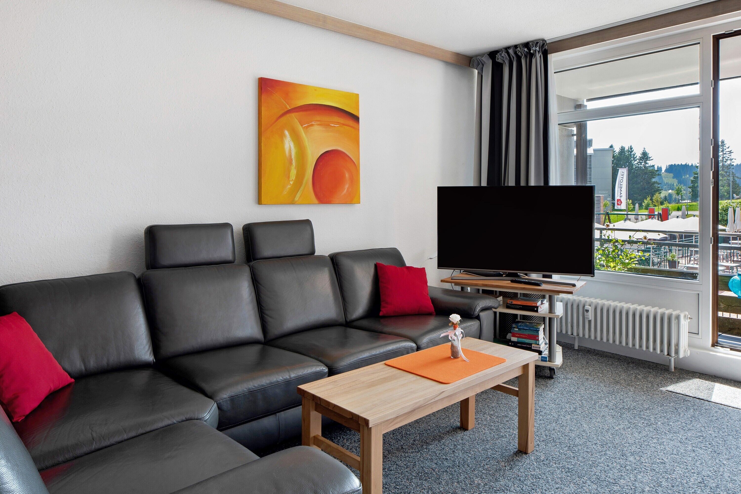 A modern hotel room with a black leather sofa, a wooden coffee table, a flat-screen TV, and a large window offering a view of the outdoors.