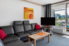 Une chambre d’hôtel moderne avec un canapé en cuir noir, une table basse en bois, un téléviseur à écran plat et une grande fenêtre donnant sur l’extérieur.