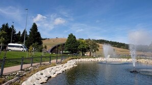 Um parque com lago, fonte, uma carrinha branca e paisagem com colinas.