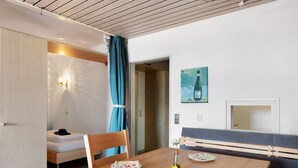 Um quarto com uma mesa de jantar de madeira, uma cama com cabeceira branca, um banco, uma luminária fixada na parede e um quadro com uma garrafa de vinho.
