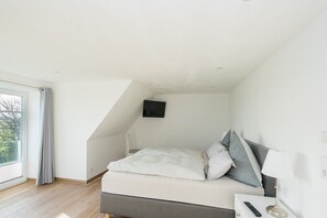 Un dormitorio con cama, televisor montado en la pared, mesita de noche con lámpara y ventana con cortinas.
