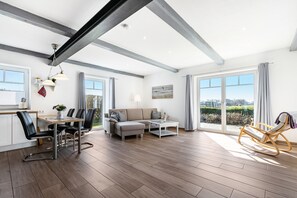 Ein großzügiger Wohnbereich mit Esstisch, Sofa und Schaukelstuhl, großen Fenstern und Blick ins Freie.