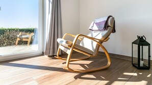 Ein Zimmer mit Schaukelstuhl, kleinem Tisch, Laterne und Fenster mit Blick auf Grünflächen.